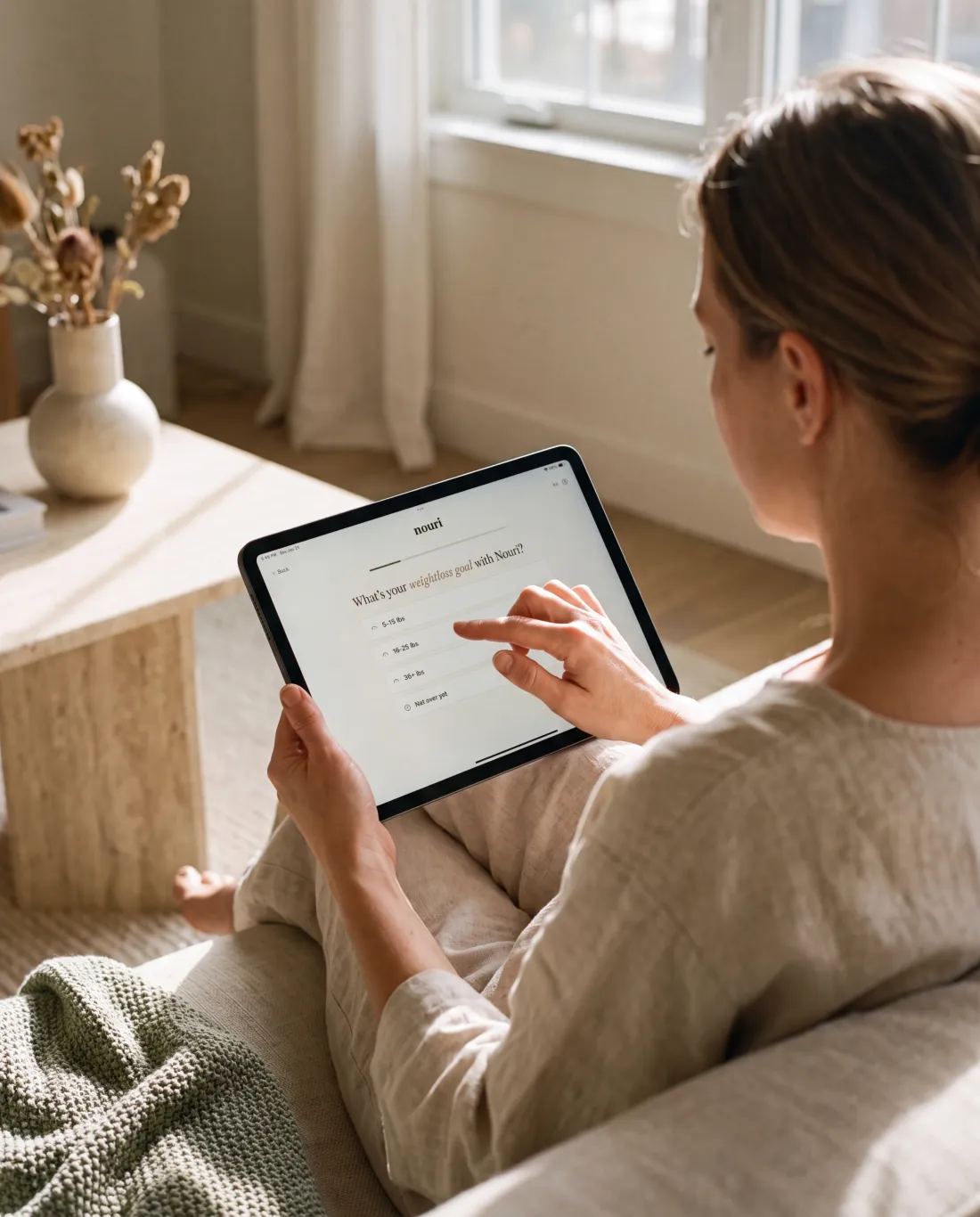 A woman on a linen sofa completes Nouri's weight-care questionnaire on a tablet at home.